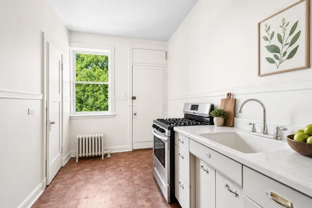 Bright kitchen with white cabinets, marble-look countertop, stainless steel gas stove, and window with view of green trees.