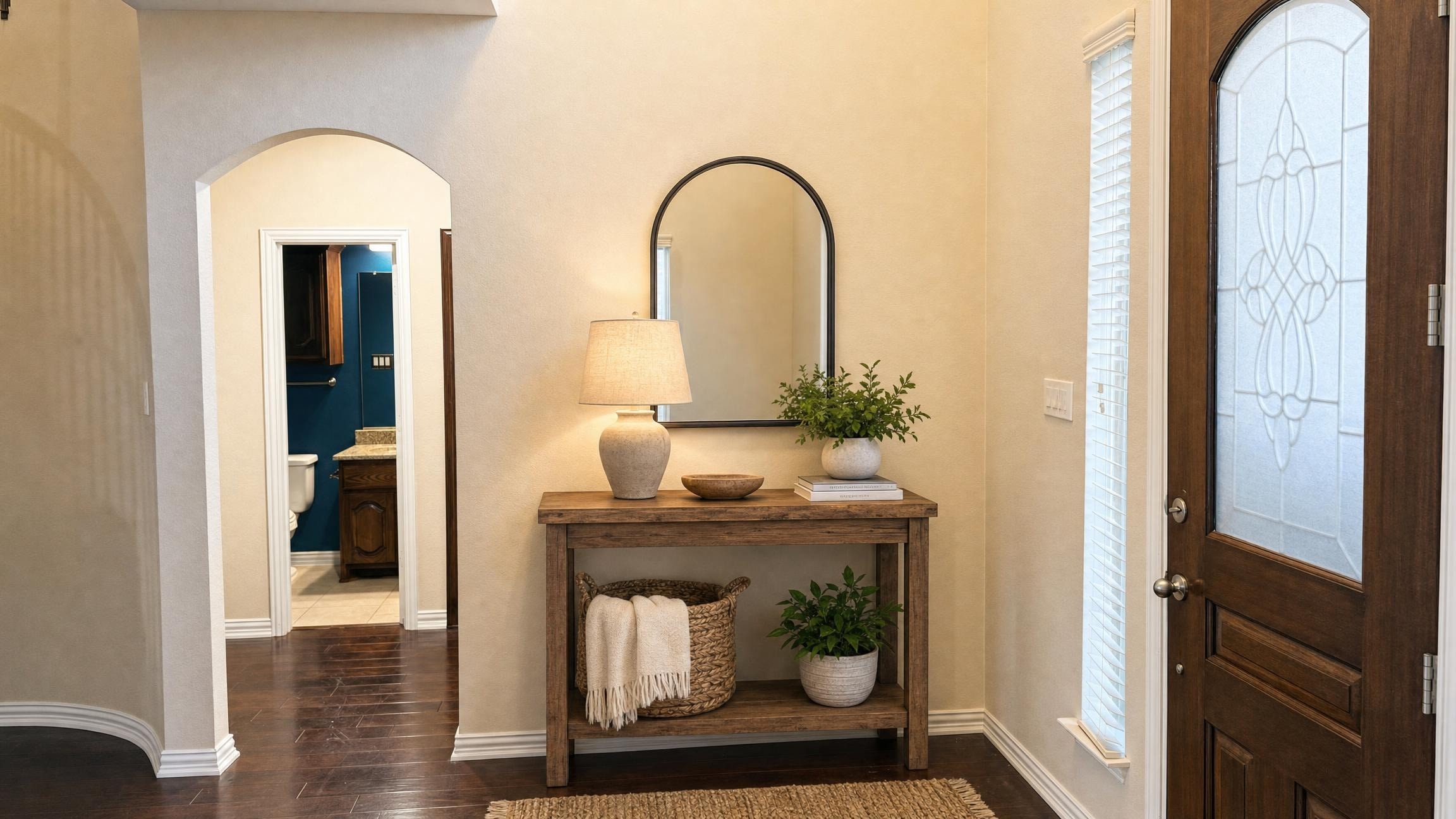 Entryway with a wooden console table topped with a lamp, decorative bowl, plant, and a basket with a throw beneath, next to a window and wooden front door.