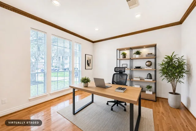 Modern home office with large windows, light wood desk with black metal legs, ergonomic black mesh chair, open black shelving, and a large potted plant.