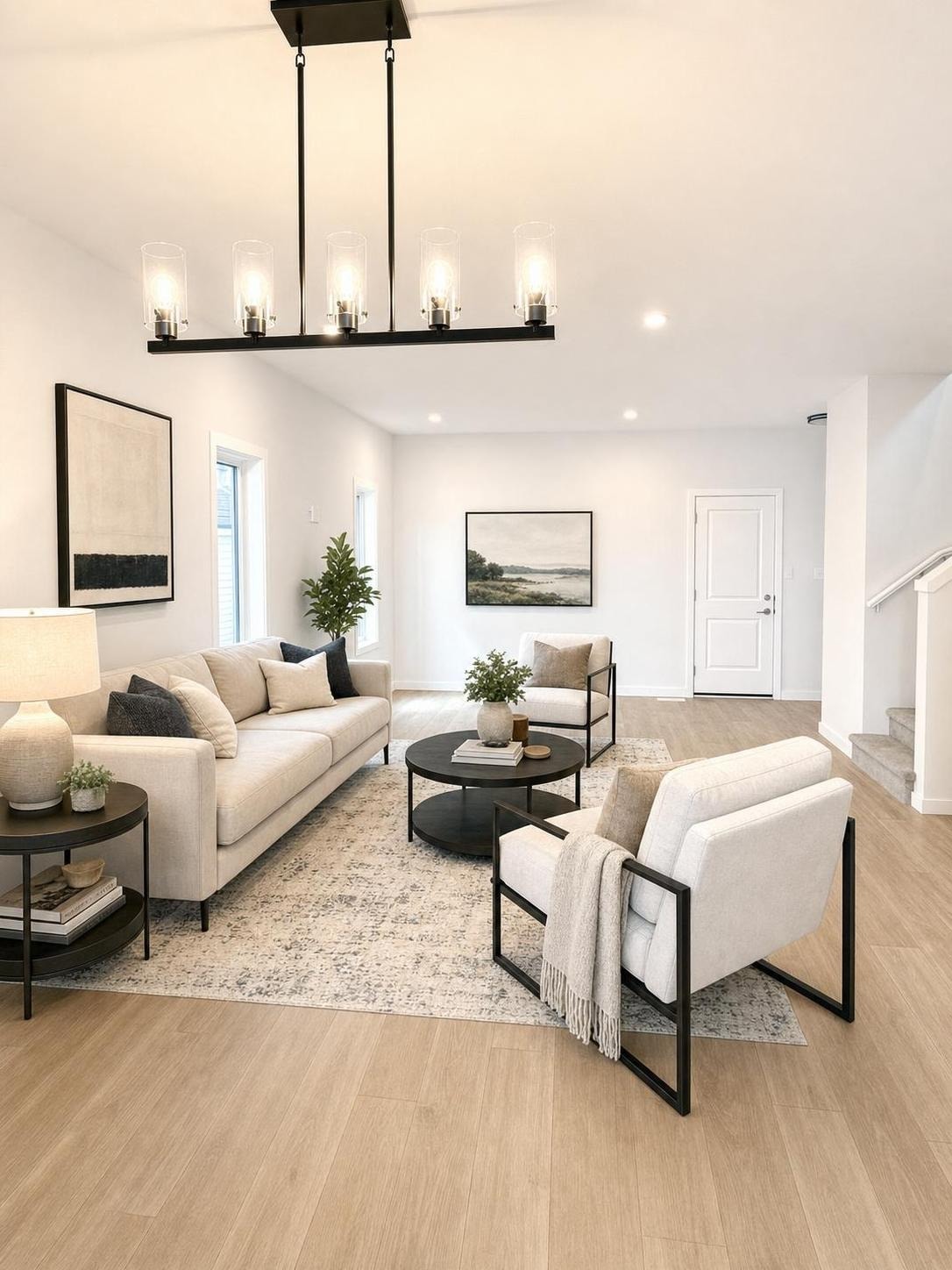 Bright modern living room with beige sofa, matching armchairs, black metal tables, light wood floor, and abstract art.