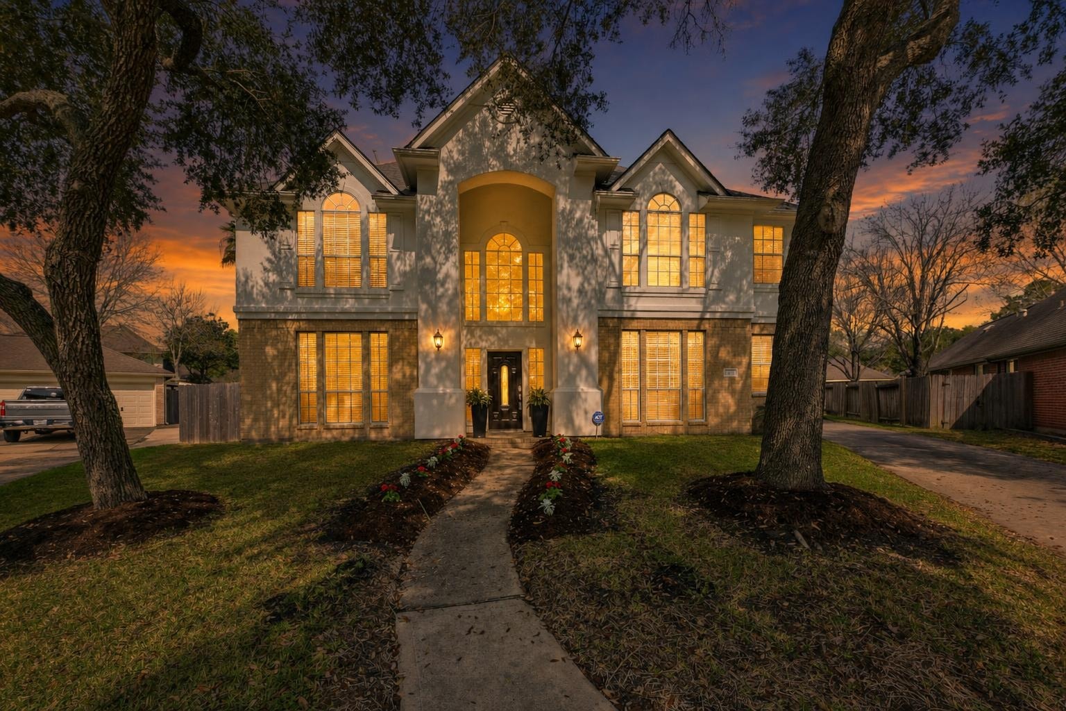 Two-story traditional house with lit windows and central arched entryway at dusk, surrounded by lawn and trees.