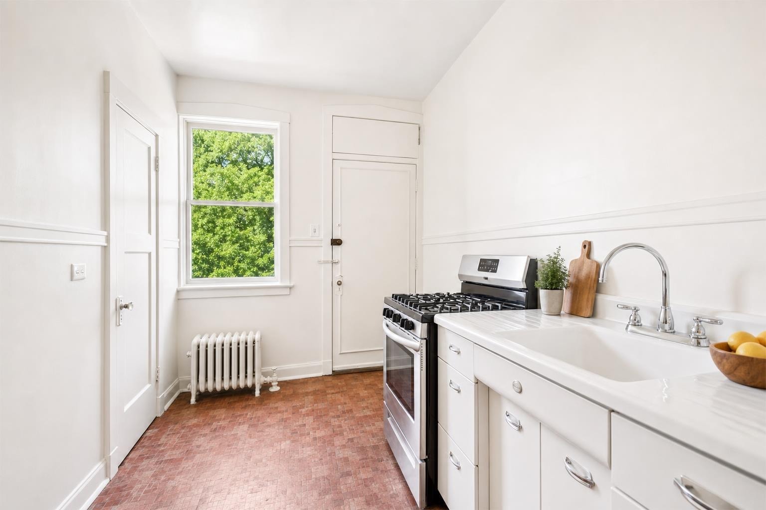 Bright kitchen with white cabinets, stainless steel gas stove, white marble-look countertop, and large window showing green trees.