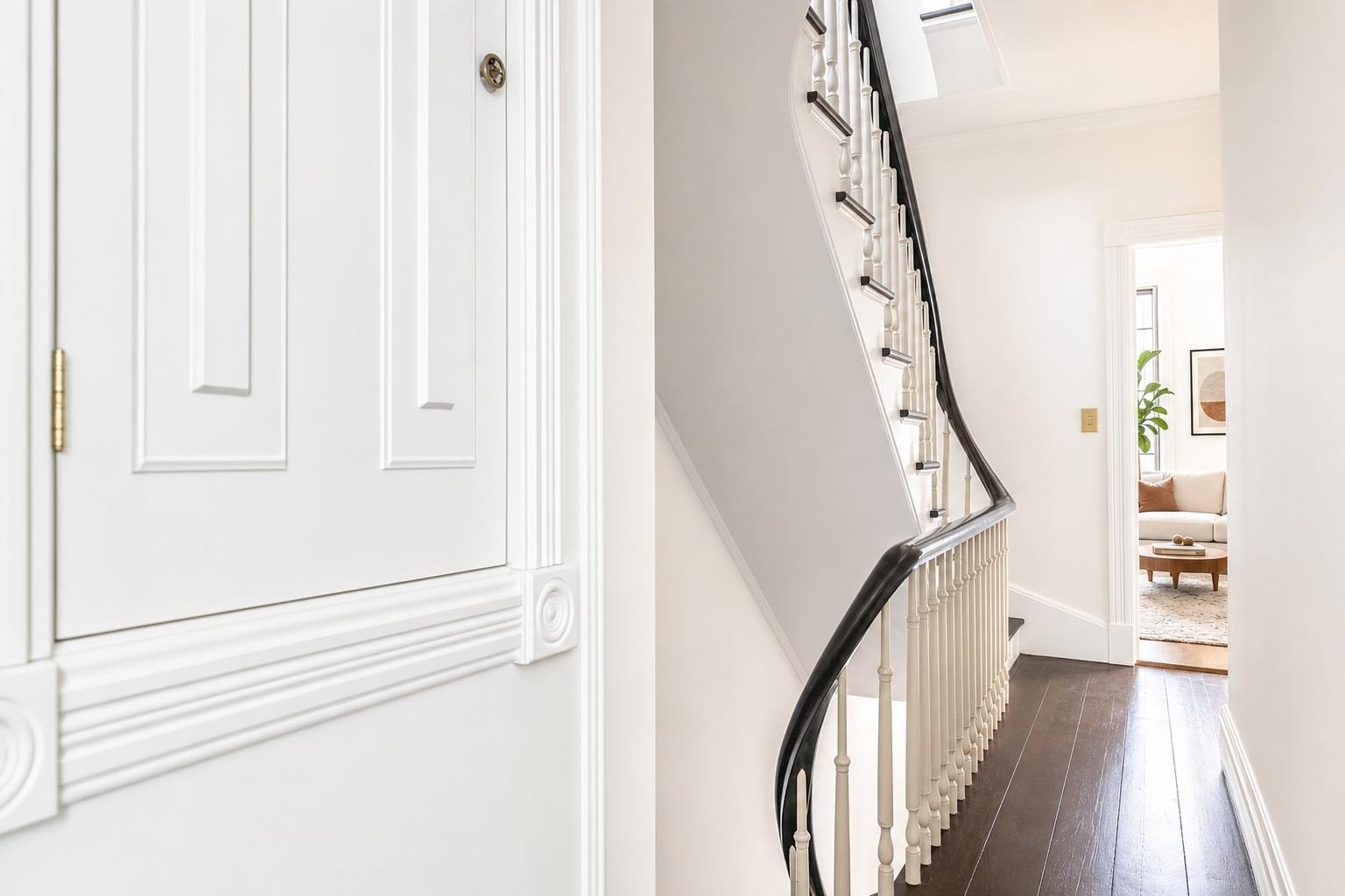 Bright hallway with white walls and black handrail staircase leading to a living room with a beige sofa and round wooden coffee table.