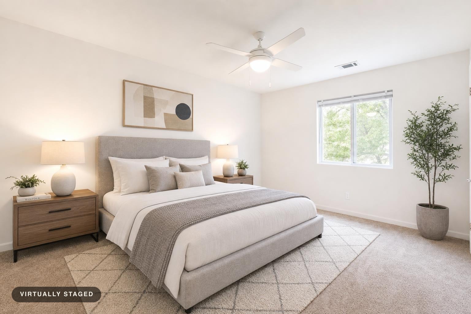 Modern bedroom with gray upholstered bed, two wooden nightstands with white lamps, a patterned rug, and a potted plant by the window.