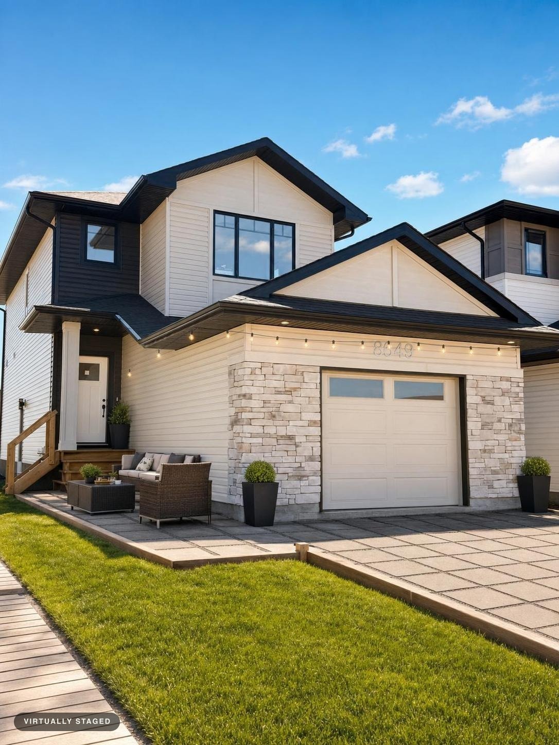 Modern two-story house with beige siding, stone accents, black-trimmed large windows, and a small outdoor patio set with greenery.