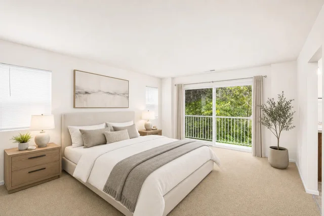Bright modern bedroom with a large upholstered bed, two wooden nightstands with lamps, and a large window with cream curtains overlooking greenery.