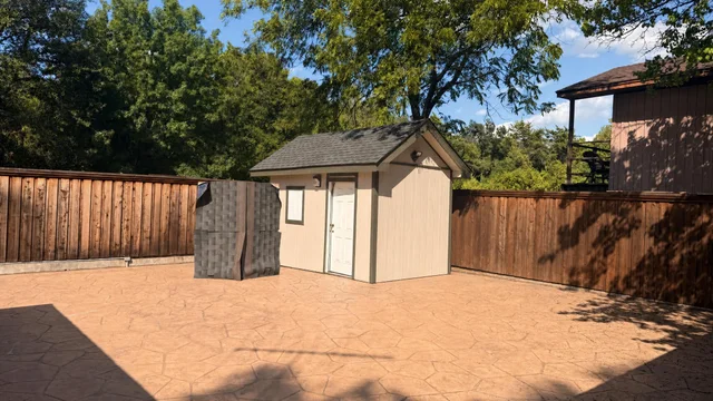 Backyard with stamped concrete floor, wooden fence, small cream-colored shed, and a dark outdoor storage box under trees.