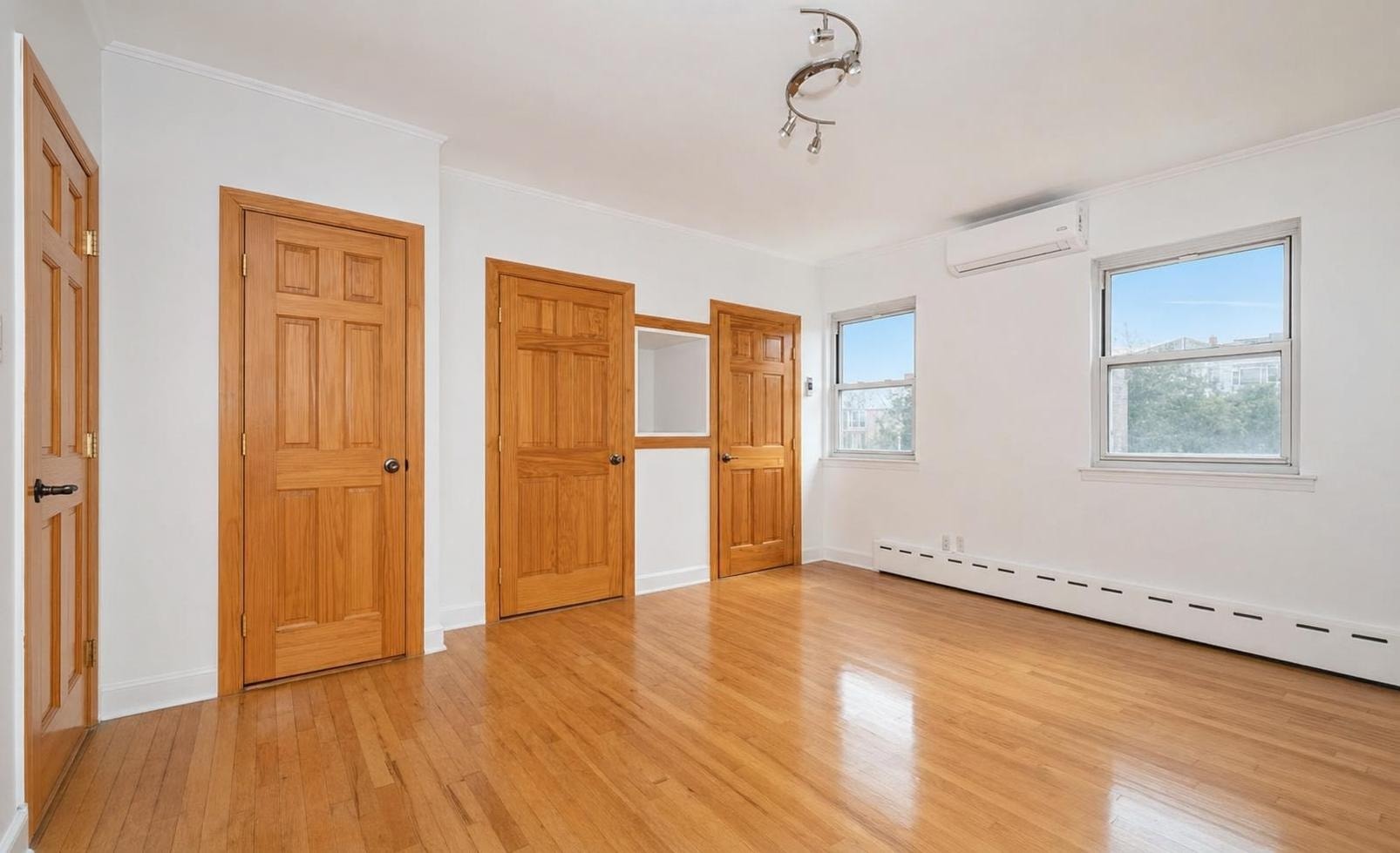 Empty living room with wooden floor, three natural wood doors, two windows, and white walls with an air conditioning unit.