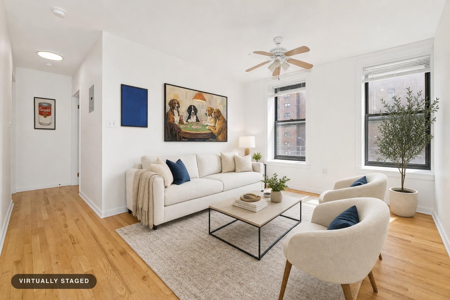 Living room with beige sofa, two cream armchairs with blue pillows, light wood floor, framed artwork, and large windows.
