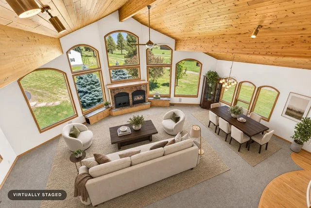 Modern living room with vaulted wood ceiling and arched windows, featuring a beige sofa, round armchairs, dark coffee table, dining set, and fireplace.