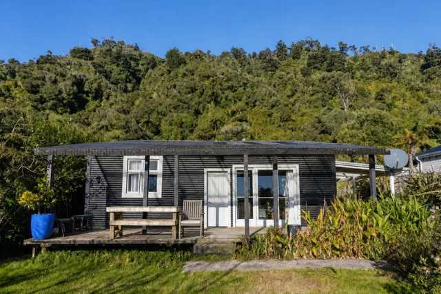 Small rustic cabin with dark wood siding, white doors and windows, a wooden deck with bench and chair, surrounded by plants and grass.