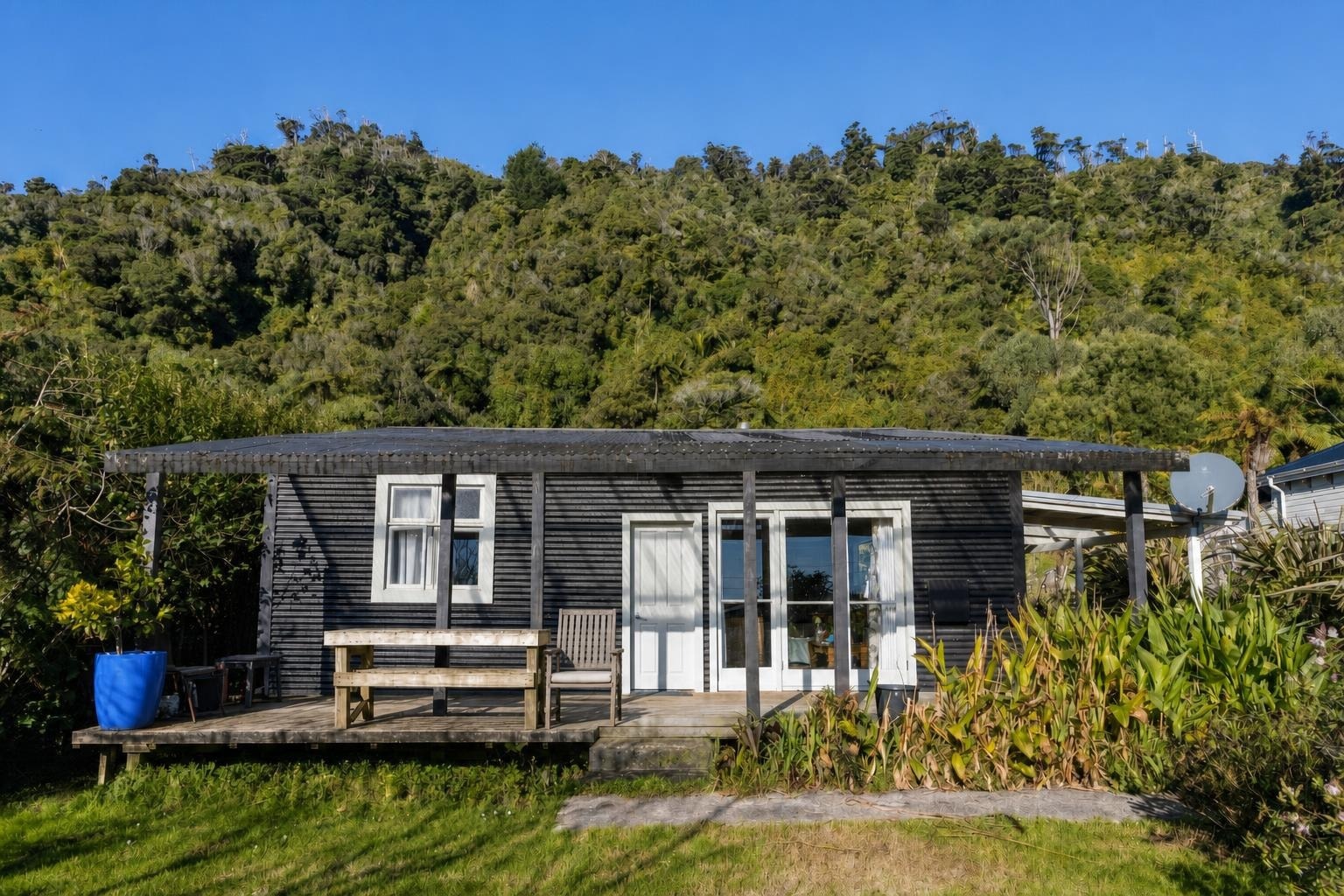 Small rustic cabin with dark wood siding, white doors and windows, a wooden deck with bench and chair, surrounded by plants and grass.