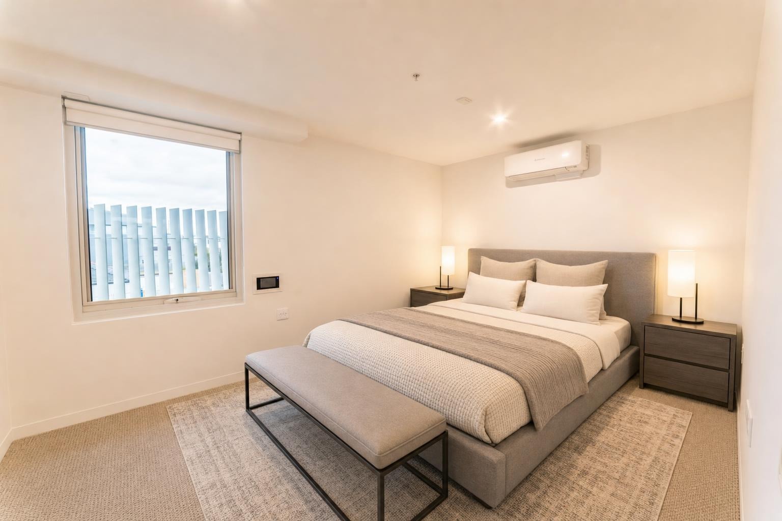 Modern bedroom with grey upholstered bed, two nightstands with lamps, a bench at foot, and a window with outdoor view.