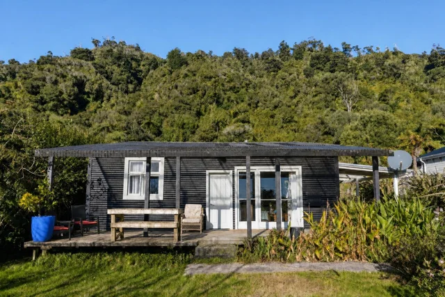 A black cabin with white windows and door on a wooden deck surrounded by lush green plants and hills under a clear blue sky.