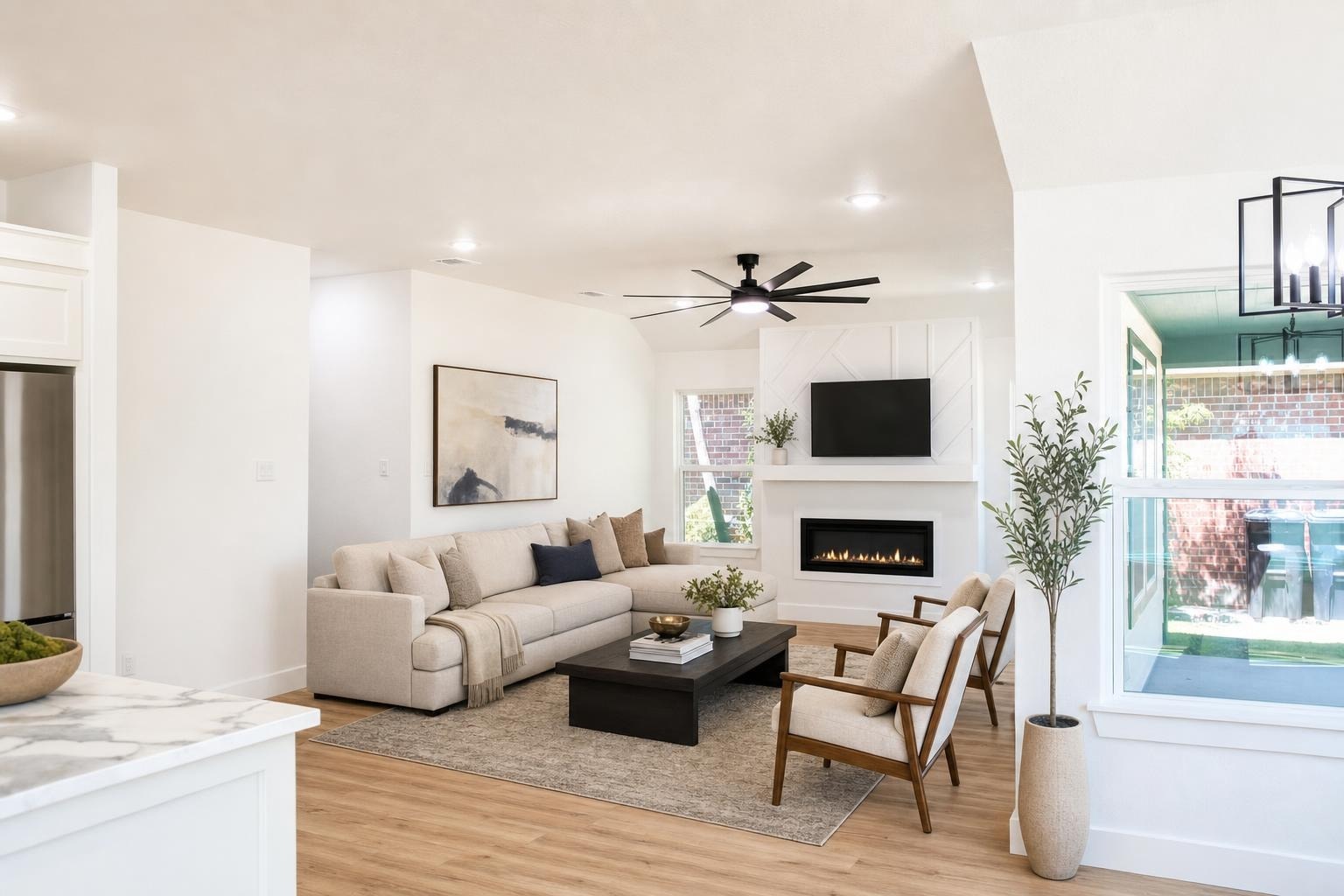 Bright modern living room with beige sectional sofa, wood armchairs, black coffee table, fireplace, and ceiling fan.