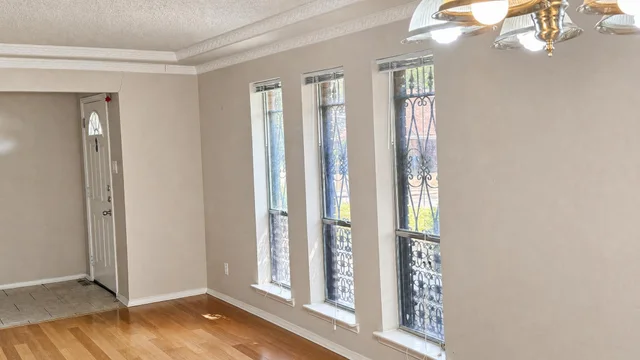 Empty living room with light hardwood floor, three tall windows with black decorative iron grilles, beige walls, and ceiling light fixture.