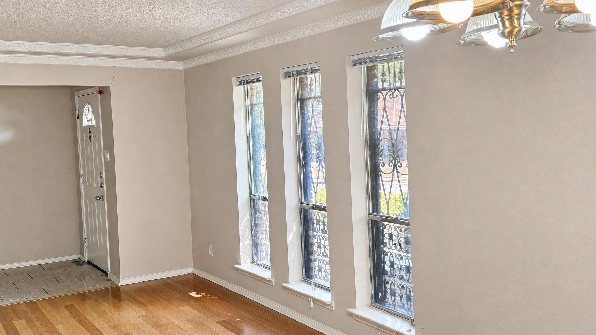 Empty living room with light hardwood floor, three tall windows with black decorative iron grilles, beige walls, and ceiling light fixture.