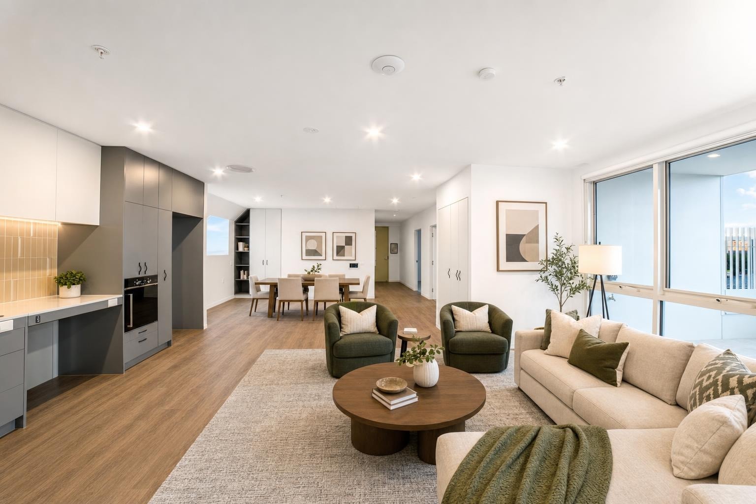 Modern living room with beige sofa, two green armchairs, round wood coffee table, and large windows.