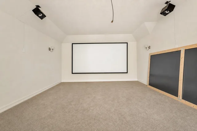 Empty living room with white walls, beige carpet, projector screen on wall, and black panel with wood frame on side.