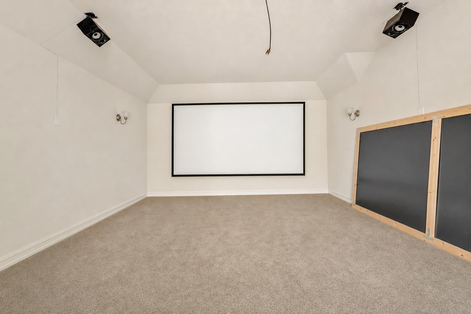 Empty living room with white walls, beige carpet, projector screen on wall, and black panel with wood frame on side.