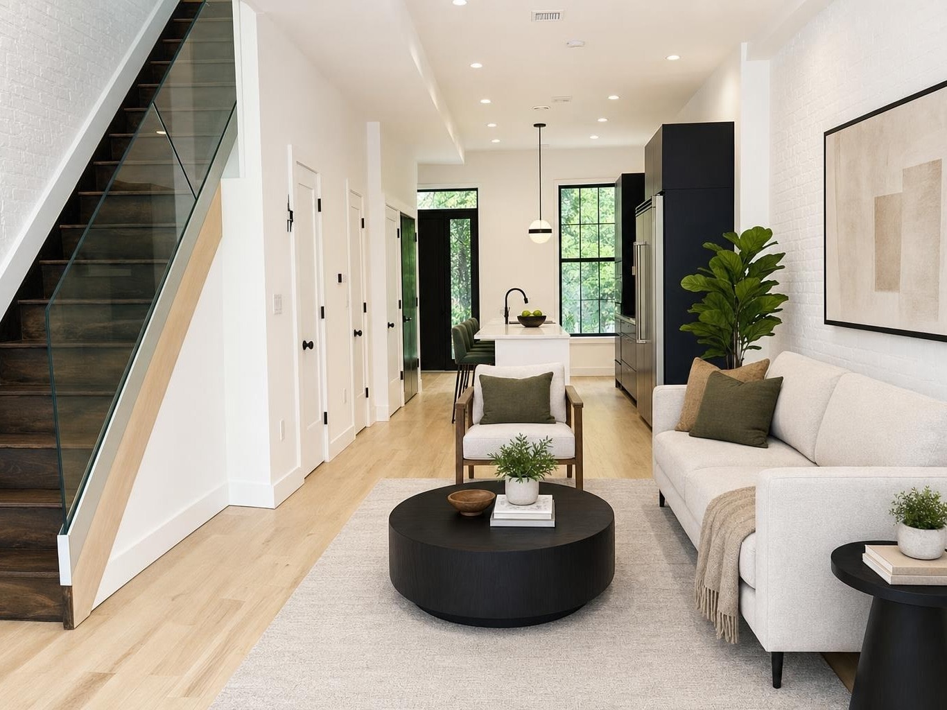 Modern living room with cream sofa, accent chair, black round coffee table, light wood floors, and stairs with glass railing.