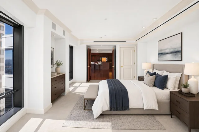 Modern luxury bedroom with beige upholstered bed, dark wood nightstands, navy accents, and large windows letting in natural light.