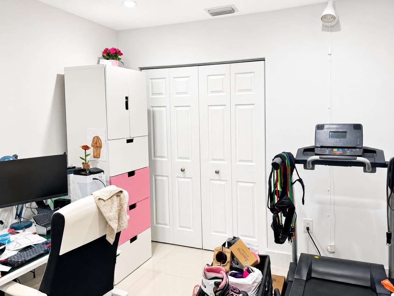 Modern home office with desk, computer, white and pink storage drawers, and a treadmill with resistance bands.
