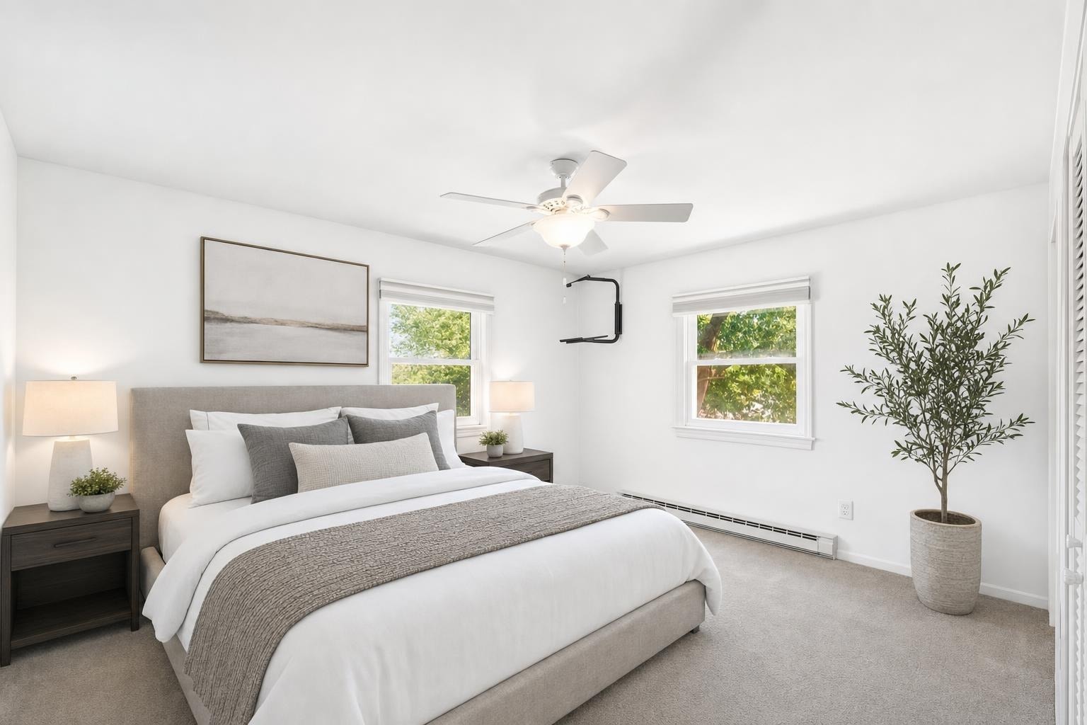 Modern bedroom with beige upholstered bed, white and gray bedding, two dark wood nightstands with lamps, a potted plant, and two windows.