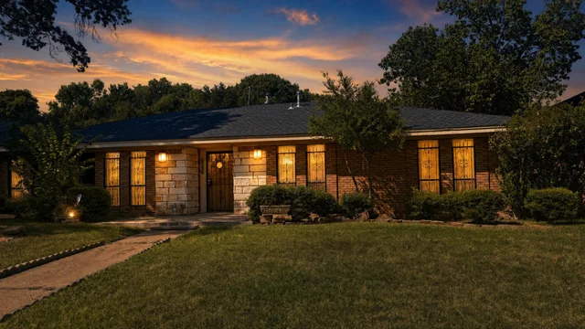 Traditional one-story brick and stone house with lit windows and front yard pathway at dusk.