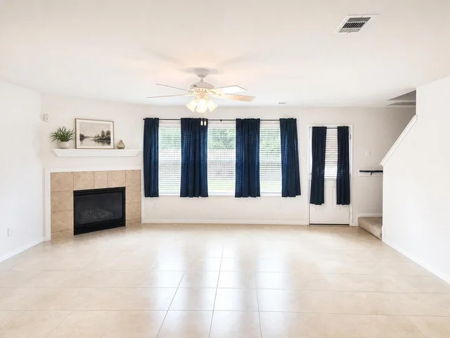 Empty living room with beige tile floor, white walls, beige tile fireplace, blue curtains on windows and door, staircase, and ceiling fan.