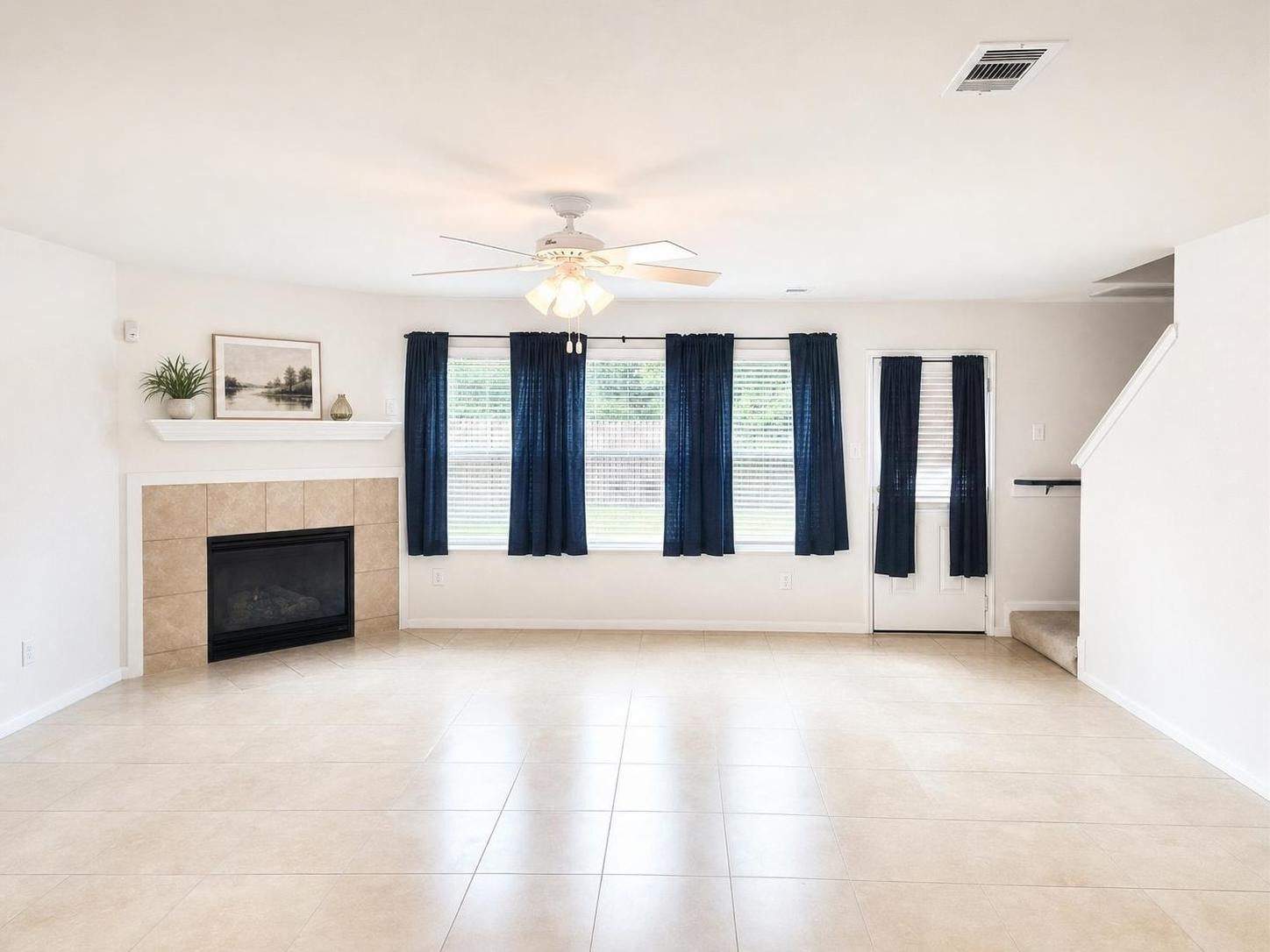 Empty living room with beige tile floor, white walls, beige tile fireplace, blue curtains on windows and door, staircase, and ceiling fan.
