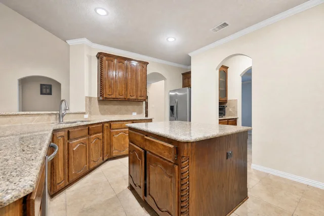 Kitchen with brown wood cabinets, granite-look countertops, central island, tiled floor, and stainless steel refrigerator.