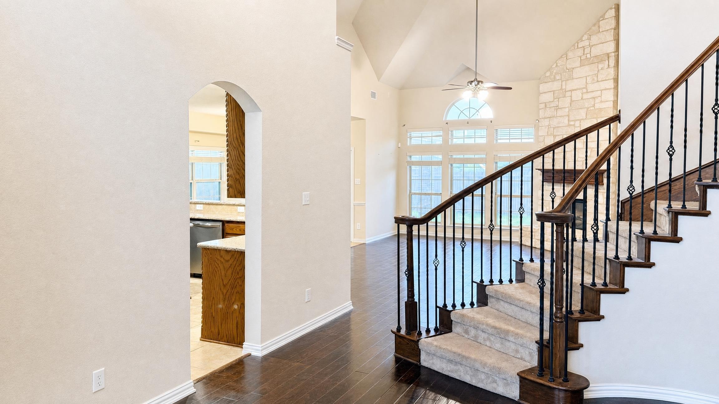 Modern living room with dark hardwood floors, white walls, vaulted ceiling, large windows, and a carpeted staircase with iron balusters.