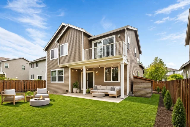 Backyard with a two-story beige house, outdoor seating with light cushions, fire pit, wooden fence, and green grass lawn.