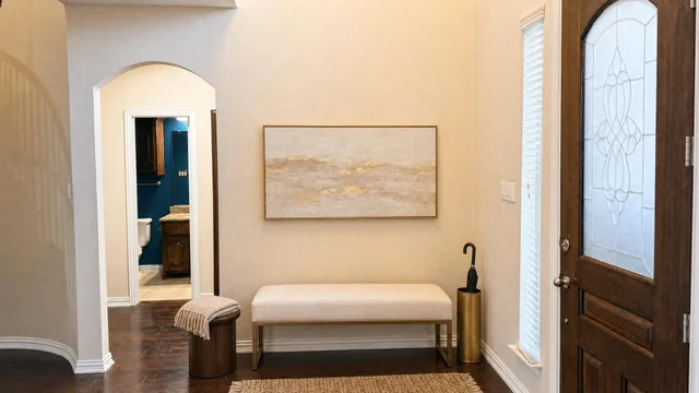 Entryway with a cream upholstered bench below gold-framed abstract art, an umbrella stand, and a wood door with decorative glass