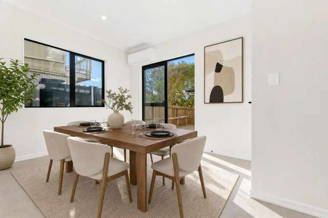 Modern dining room with rectangular wooden table, six beige upholstered chairs, abstract wall art, and large windows.