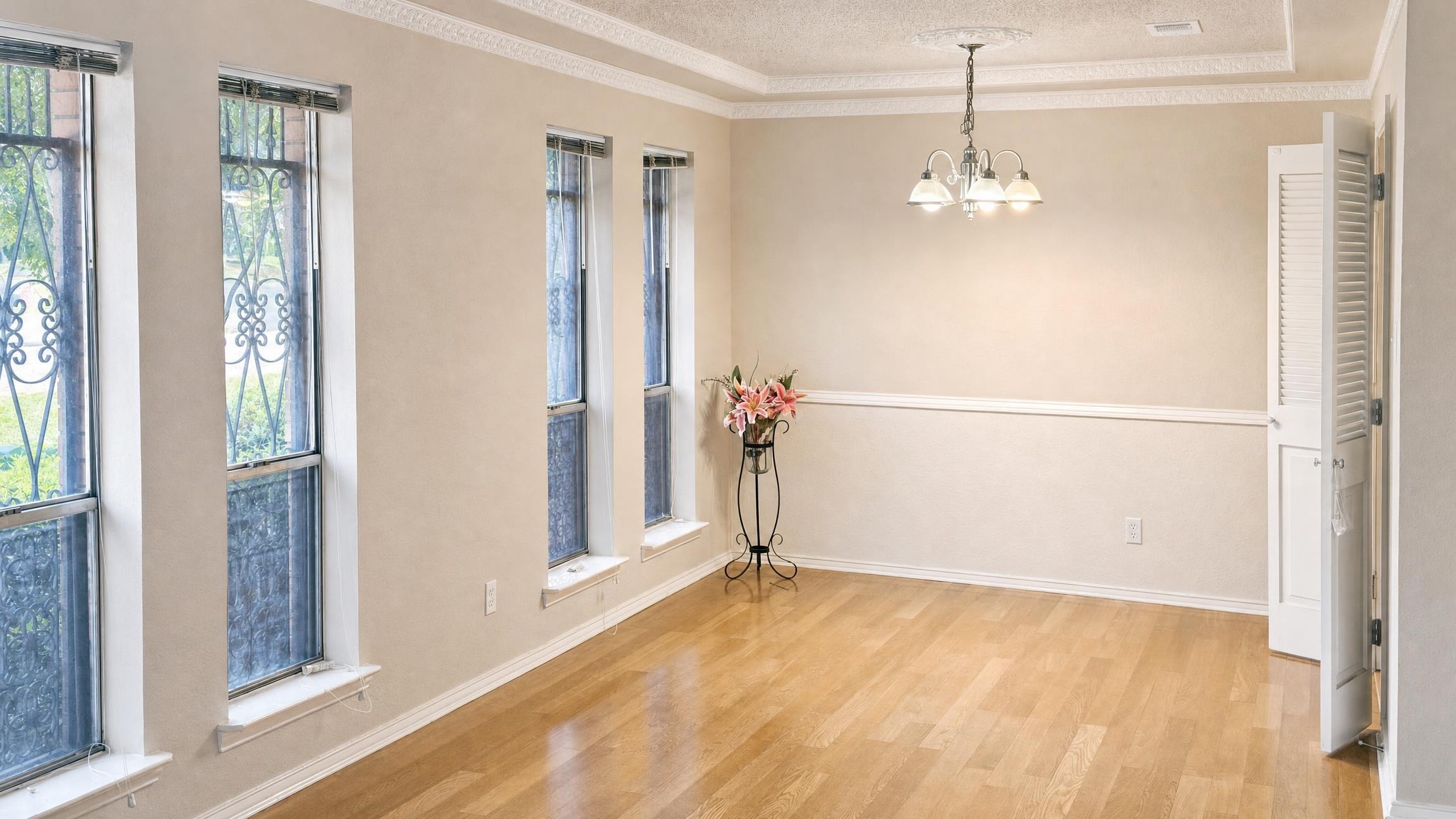 Empty dining room with beige walls, three tall windows with iron grilles, wood floor, and a small chandelier.