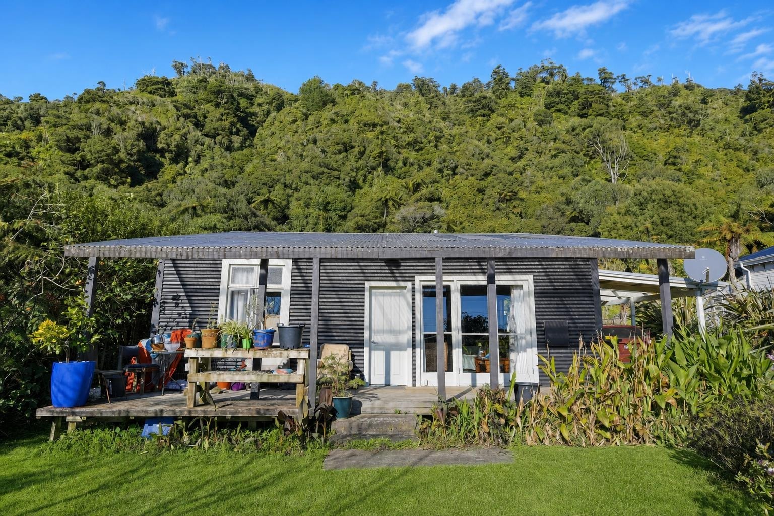 House exterior with covered porch, potted plants on a wooden deck, and lush green hill in the background under blue sky.