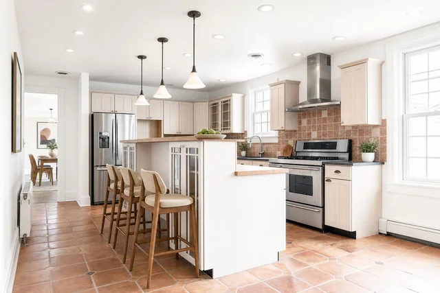 Bright kitchen with light wood cabinets, stainless steel appliances, wood-tone bar stools at center island, and terracotta floor tiles.