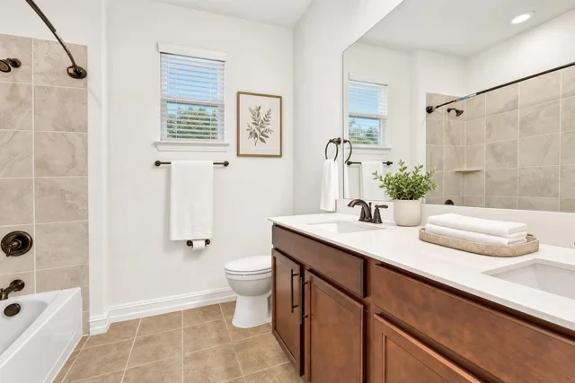 Modern bathroom with beige floor tiles, dark wood vanity with two sinks, white towels, and a tiled bathtub with bronze fixtures.