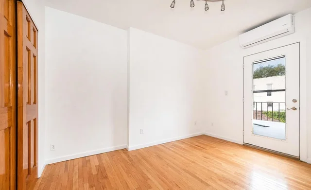 Empty living room with hardwood floors, white walls, a wooden closet door on left, and a glass door on the right.