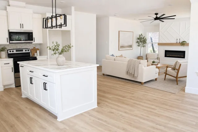 Bright living room with white kitchen island, light wood floor, beige sofas, wooden armchair, and black ceiling fan.