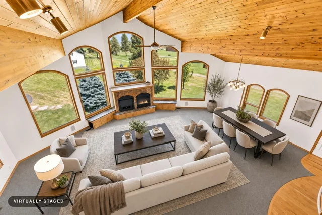 Open living and dining room with vaulted wood ceiling, large arched windows, beige sectional sofa, armchair, dark wood coffee table, and dining set.
