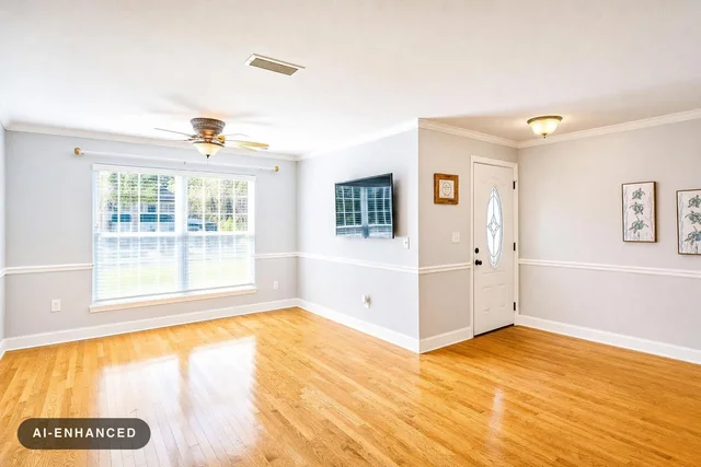Empty modern living room with hardwood floor, ceiling fan, large window with blinds, wall-mounted TV, and a white door with glass panel.