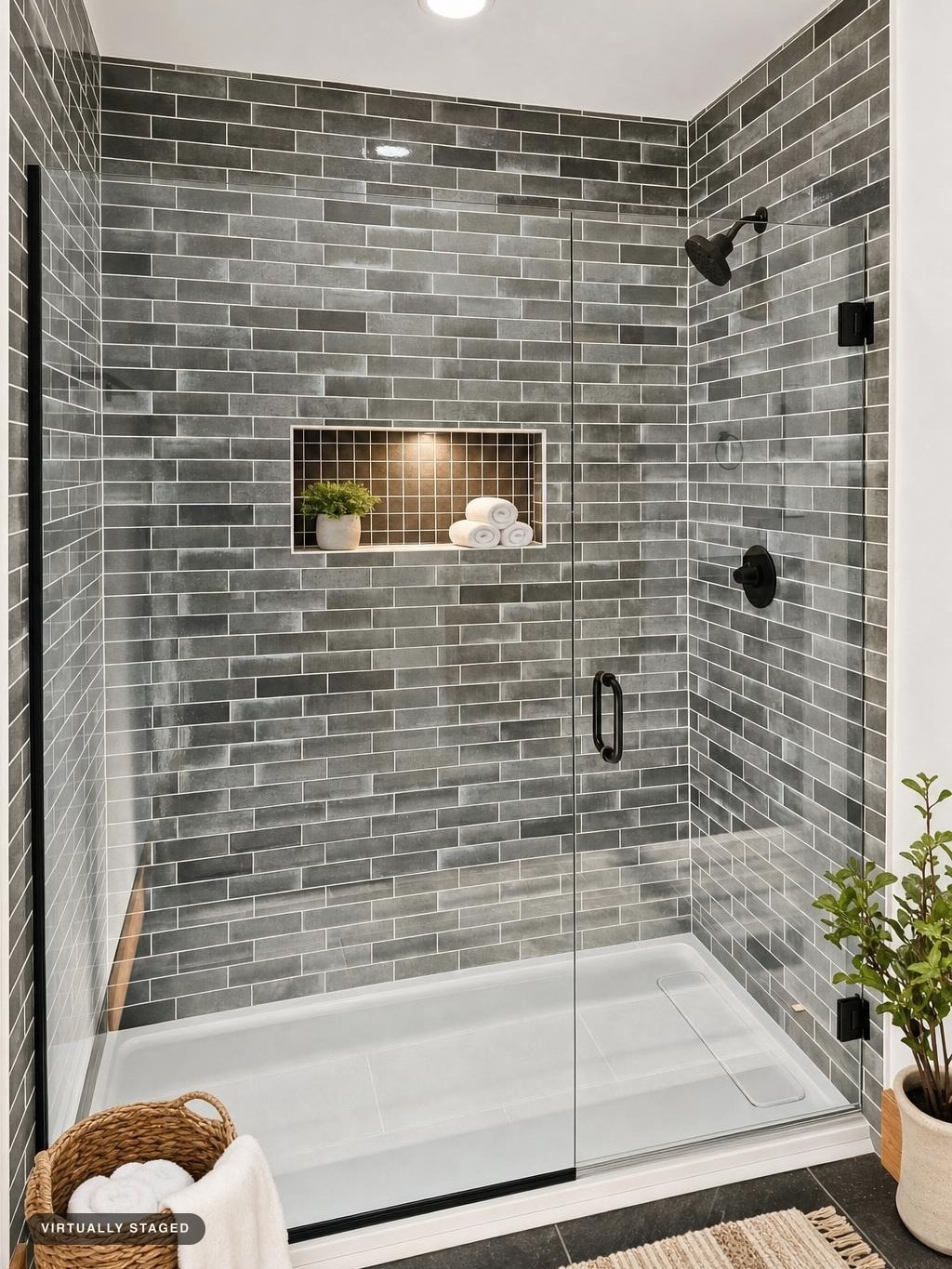 Glass-enclosed shower with gray subway tiles, built-in niche with lighting, black fixtures, and a woven basket with towels.