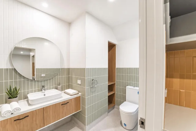 Modern bathroom with floating vanity, oval mirror, towel rolls, and half olive green tiled walls with wood open shelves.