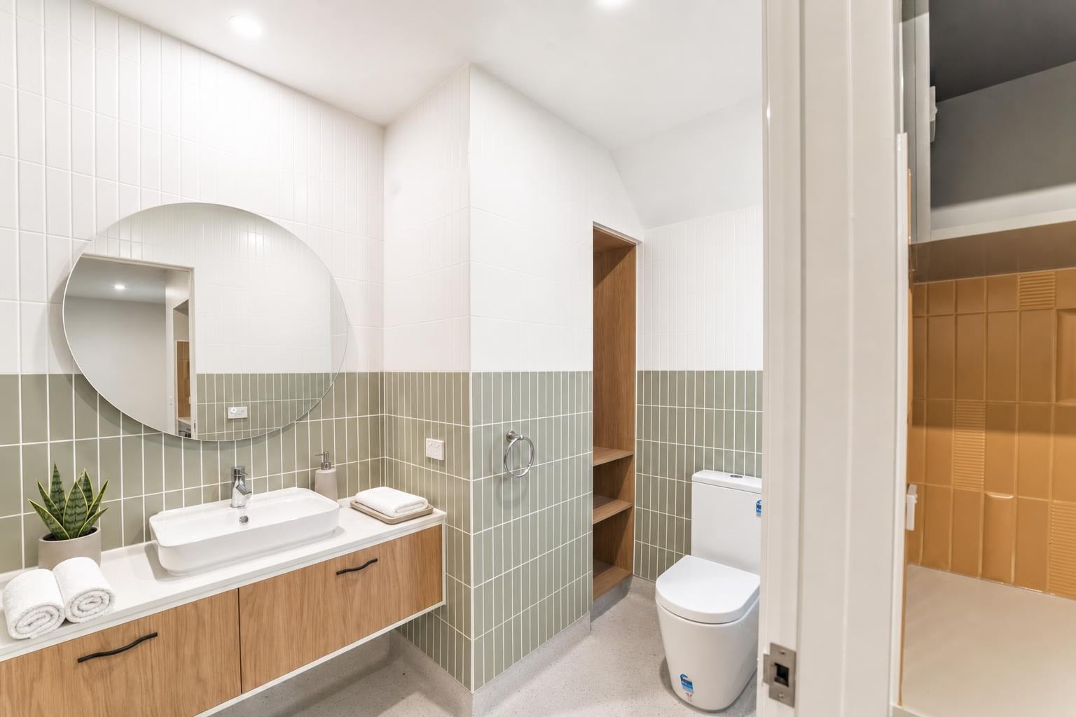 Modern bathroom with floating vanity, oval mirror, towel rolls, and half olive green tiled walls with wood open shelves.