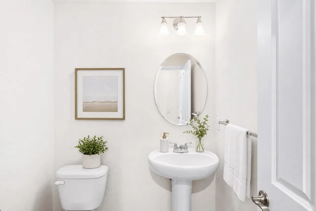 Small bathroom with white pedestal sink, oval mirror, a picture with light wood frame, toilet with plant, and white towel on metal rack.