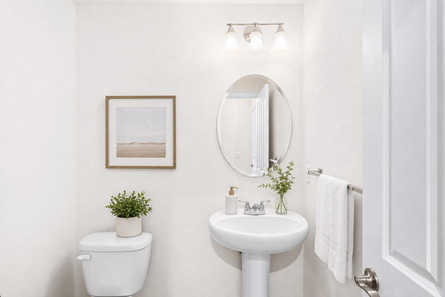 Small bathroom with white pedestal sink, oval mirror, a picture with light wood frame, toilet with plant, and white towel on metal rack.