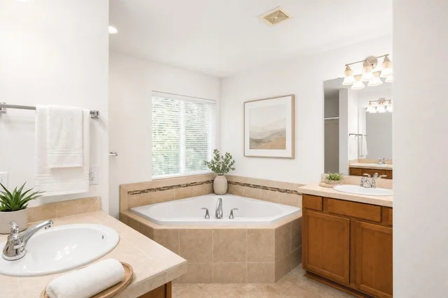 Bright bathroom with corner bathtub, wood-tone vanity, white sink, beige tiles, green plants, and framed wall art.